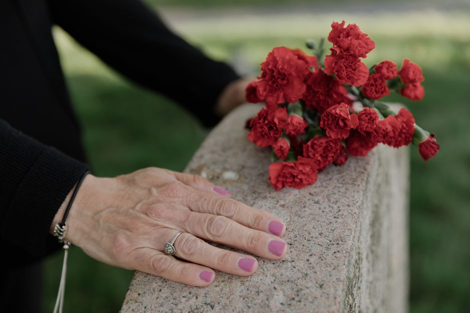 A pair of hands with pink nail polish resting on a stone ledge beside a bouquet of bright red carnations, arranged with green stems and leaves, wrapped in clear plastic, in an outdoor setting with gre