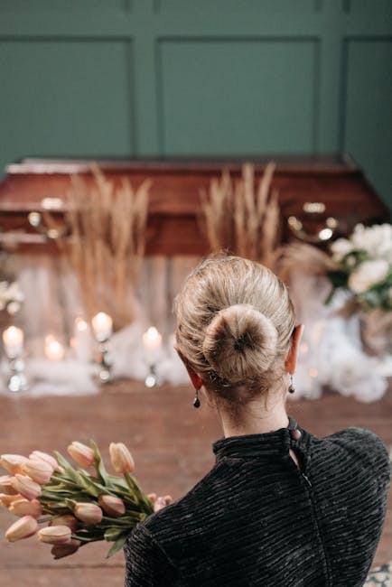 A woman with her blonde hair styled in a neat bun, wearing a dark textured jacket with a visible zipper at the back, is holding a bouquet of pale pink tulips with green stems and leaves. She is standi
