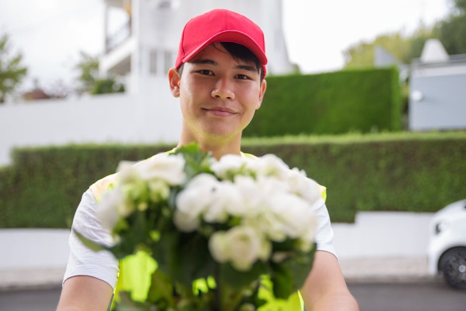 A young male flower delivery courier wearing a red cap and a high-visibility yellow vest is smiling and holding out a bouquet of fresh white roses and greenery, wrapped in clear cellophane. The bouque