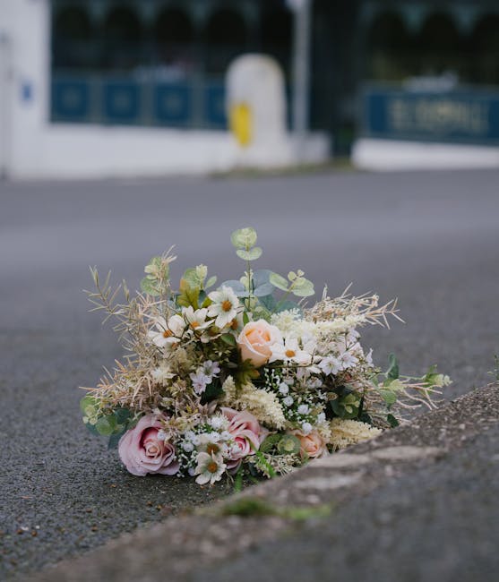A delicate floral bouquet resting on a paved surface outside, featuring soft pink roses, white daisies, and sprigs of greenery including eucalyptus and dried foliage. The arrangement is loosely gather