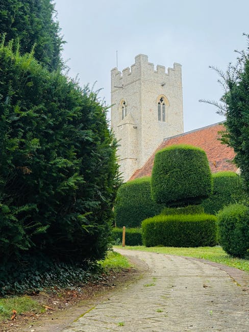 A cobbled pathway winding through a well-manicured garden with lush greenery and neatly trimmed, rounded bushes leading towards a historic stone church tower in the background. The garden is vibrant w