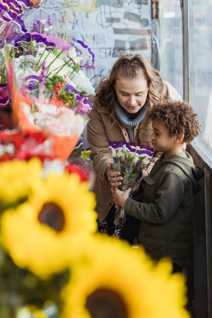 A young woman and a small girl are standing inside a flower shop near a large window, engaged in a moment of exchange. The woman, with curly blonde hair and wearing a beige coat, hands a bouquet of pu